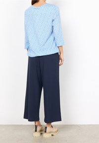 Top azzurro chiaro con motivo astratto bianco, maniche 3/4; pantaloni a gamba larga blu navy; scarpe beige slip-on con dettagli neri a contrasto.