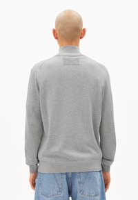 Grauer Pullover mit hohem Kragen, gerippten Bündchen und Saum sowie einem strukturierten Einsatz auf der Rückseite, getragen über hellblauer Jeans.