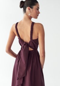 Bordo obleka z debelimi, nagubanimi naramnicami, odprtim hrbtom in zavezanim detajlom v pasu. Narejena iz mehkega materiala s tekočo silhueto.