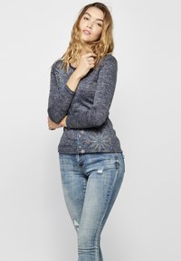 Jūrų laivyno ilgos rankovės marškiniai su sudėtingu šviesiai mėlynos ir baltos spalvos siuvinėjimu apatinėje dalyje, derinami su prakaituotais šviesiai mėlynais džinsais.