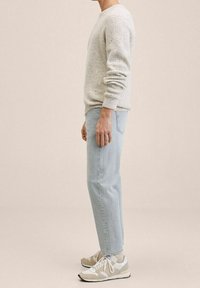 Uomo in piedi con maglione leggero lavorato a maglia, jeans azzurri e sneakers beige, su sfondo beige uniforme, mostrato di profilo dalla spalla in giù.