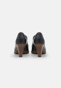 Zapatos de tacón alto de cuero negro con punta redondeada y tacón de madera de color marrón claro, que exhiben una textura suave y detalles de diseño minimalistas.
