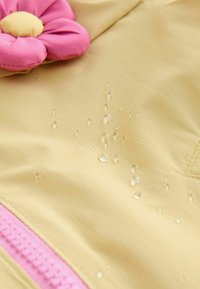 Tela impermeable amarilla con decoración de flores rosas; gotas de agua en la superficie, mostrando las propiedades hidrófugas del material.