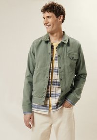 Groene canvasjas met drukknoopsluiting, voorzak, gedragen over een geruit shirt en een gele T-shirt. Crème-kleurige broek maakt de look compleet.