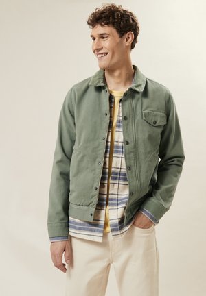 Green canvas jacket with snap buttons, front pocket, worn over a plaid shirt and yellow tee. Cream-colored pants complete the look.