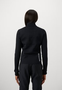 Pessoa de costas para a câmara, vestindo uma blusa preta de manga longa justa e calças pretas, contra um fundo claro e simples.