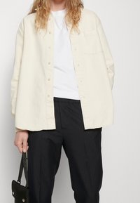 Camicia button-up di un beige chiaro, con una vestibilità rilassata, taschino sul petto e texture morbida, abbinata a pantaloni scuri a righe e a una borsa piccola.