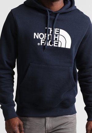 Osoba nosząca ciemnoniebieską bluzę z kapturem z przednią kieszenią i białym logo "The North Face" na piersi.