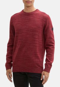 Pull en tricot rouge avec un col rond et des longues manches. Présente un motif texturé et un petit logo noir sur la manche gauche.
