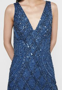 Bezramínkové modré šaty s hlubokým výstřihem do V, ozdobené flitry a geometrickým vzorem, s texturovanou tkaninou a priléhavým střihem.