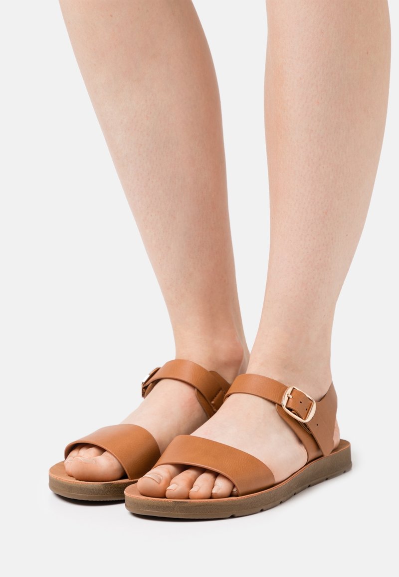 Pieds portant des sandales en cuir marron avec des brides autour de la cheville et des boucles, présentés sur un fond clair.