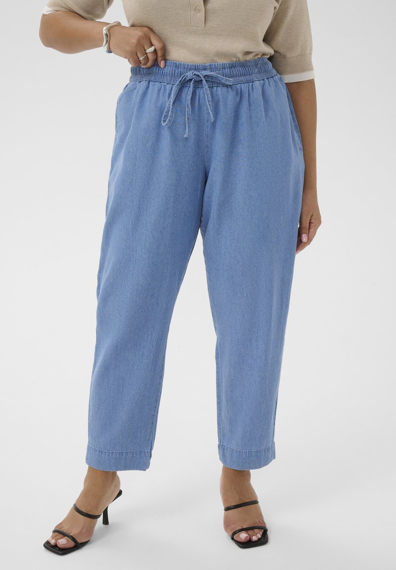 Femme portant un pantalon bleu clair à cordon, un haut beige à manches courtes, des sandales noires à talons, avec des bagues et un bracelet à la main gauche.