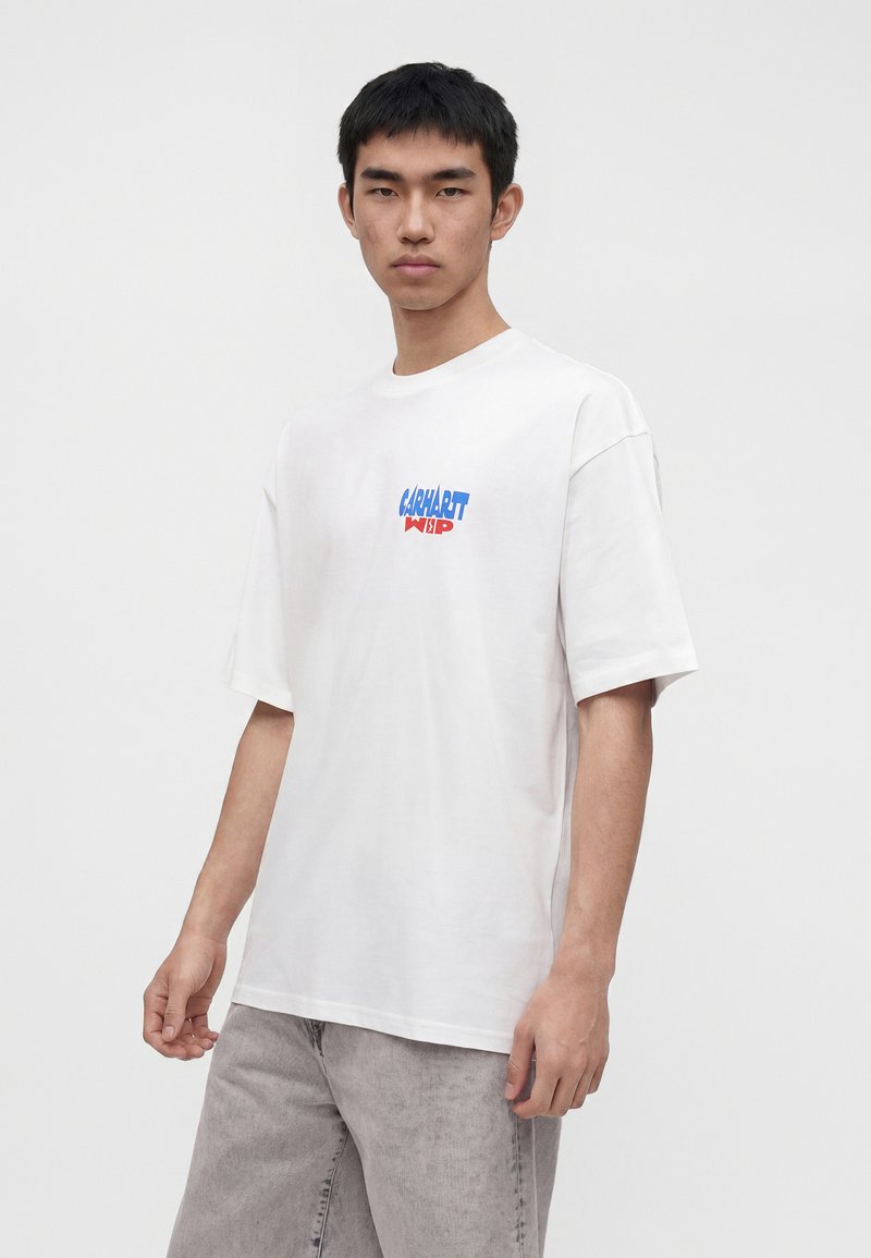 Giovane uomo che indossa una T-shirt bianca oversize con il logo blu e rosso "Carhartt WIP", abbinata a jeans grigio chiaro, in piedi davanti a uno sfondo uniforme.
