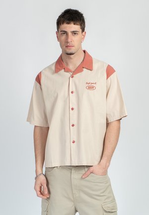 Ung mann iført beige kortermet skjorte med rustfarget krage og skulderdetaljer, stående med den ene hånden i lommen.