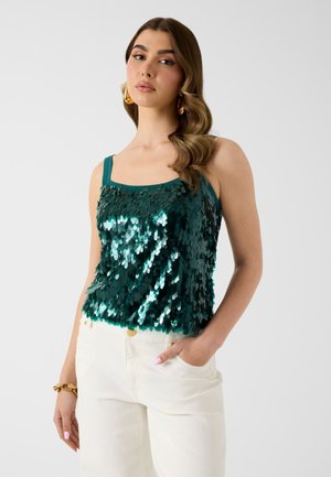 Donna con capelli mossi e riga laterale che indossa grandi orecchini d'oro, un top verde senza maniche con paillettes, pantaloni bianchi e un braccialetto d'oro al polso sinistro.