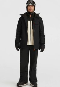 Must have veekindel must suusajakk kreemika paneeliga, lukuga tasku, millele on sobitatud mustad lumepüksid ja kindad, kantud kiivri ja prillidega.