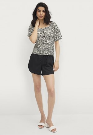 Schwarzes und weißes gepunktetes kurzärmeliges Oberteil, kombiniert mit schwarzen Shorts. Das Oberteil hat einen quadratischen Ausschnitt und geraffte Ärmel aus glattem Stoff.