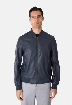 Uomo che indossa una giacca bomber blu navy con zip e tasche con zip e pantaloni grigio chiaro, in piedi davanti a uno sfondo chiaro e semplice.