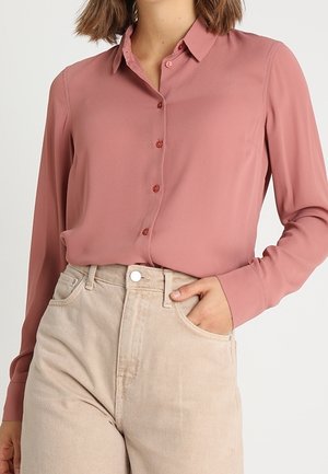 Femme portant une chemise boutonnée rose poudré à manches longues rentrée dans un pantalon beige taille haute, avec une main dans la poche.
