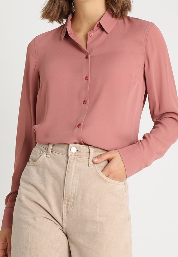 Femme portant une chemise boutonnée rose poudré à manches longues rentrée dans un pantalon beige taille haute, avec une main dans la poche.