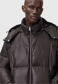 Mand iført en sort dunjakke med høj krave og en delvist løs hætte foran en ensfarvet lys baggrund.