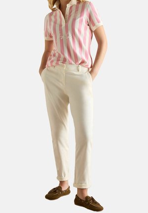 Personne portant un polo à manches courtes rayé rose et blanc, un pantalon crème retroussé et des mocassins marron, mains dans les poches.