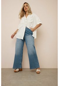 Vit skjorta med vertikala ränder, två framlommor, i kombination med vida blå jeans och träplattformsandaler.