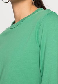 Maglietta verde a maniche lunghe in morbido tessuto, con scollo rotondo e una leggera texture, vista di lato, che mette in evidenza il colore.