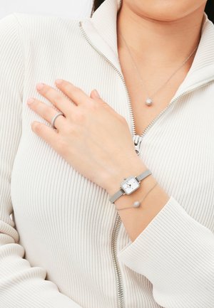Orologio da polso in rete argentata con quadrante quadrato. Accompagnato da un delicato bracciale in argento con un accento sferico. Top in crema con texture.