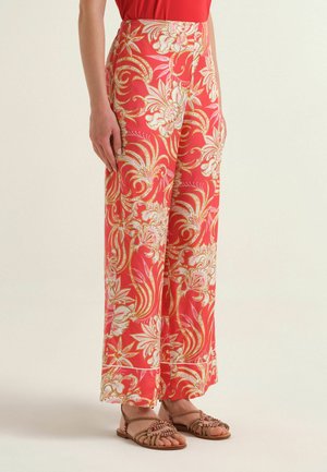 Bright coral wide-leg pants with intricate white and gold floral and swirl patterns worn with strappy brown sandals.