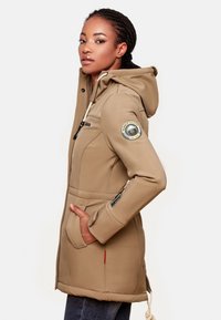 Beige hooded jacket with a zip closure, side pockets, and logo patch on the sleeve. Features drawstrings and a soft, textured fabric.
