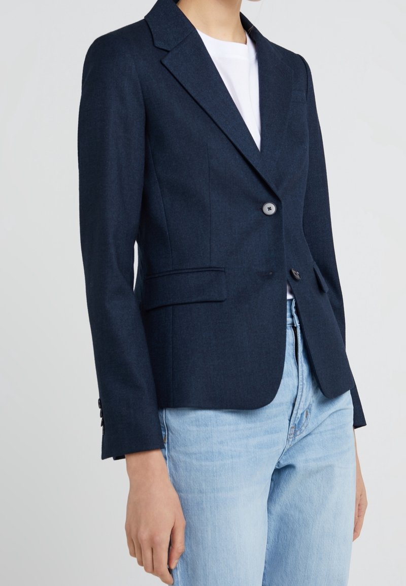 Personne portant un blazer bleu marine foncé sur un top blanc assorti à un jean bleu clair, montrée des épaules aux hanches sur un fond uni.