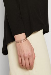 Silver metal cuff bracelet with three intertwining bands, worn on a hand. Background features a black shirt and cream-colored fabric.