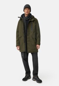 Olivgrüne Parkajacke mit Frontreißverschluss, großen Taschen und abnehmbarem Kapuze; kombiniert mit einem grauen Schal, schwarzem Pullover und dunklen Jeans.