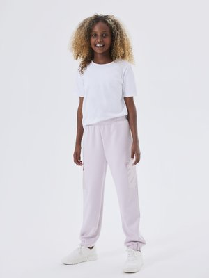 T-shirt bianco a maniche corte abbinato a pantaloni della tuta viola chiaro con tasche laterali, polsini elasticizzati e sneakers bianche su uno sfondo neutro.