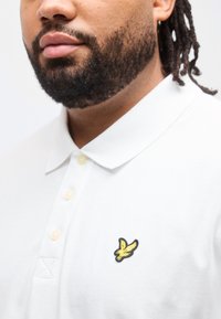 Άνδρας με μούσι και dreadlocks που φοράει λευκό πόλο μπλουζάκι με ένα μικρό κίτρινο πουλί λογότυπο στο αριστερό στήθος.