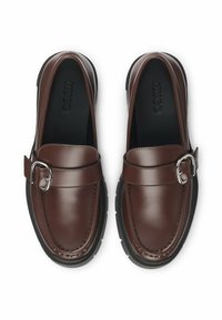 Mocassins en cuir marron avec un détail boucle argentée, bout arrondi et semelle en caoutchouc noire à finition texturée. Logo visible à l'intérieur.
