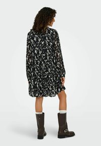 Robe à motifs noir et blanc avec des manches longues et un ourlet volants. Le mannequin porte des bottes marron montantes avec des chaussettes blanches.