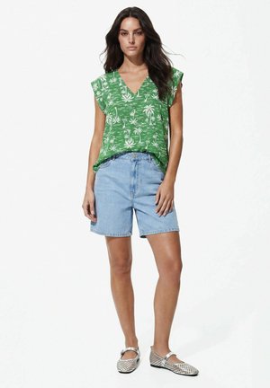 Donna con una blusa verde a maniche corte con stampa tropicale, shorts di jeans azzurri e scarpe basse bianche in mesh, in piedi davanti a uno sfondo semplice.