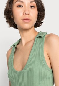 Top de tanque verde com relevos, decote profundo e tecido suave e texturizado. Apresenta detalhes arredondados nas mangas curtas com costura visível.