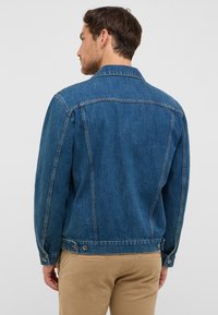 Jeansowa kurtka w średnim odcieniu niebieskiego o klasycznym kroju, z zaostrzonym kołnierzem, mankietami na guziki i regulowanym pasem. Widok z tyłu, gładka faktura.