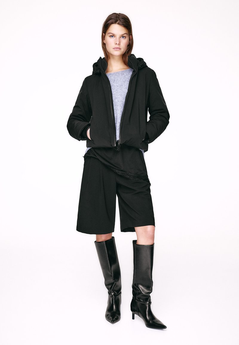 Crna dukserica s patentnim zatvaračem i kapuljačom sa užetom, siva majica ispod, crne široke kratke hlače i sjajne crne čizme preko koljena na tanke visoke potpetice.