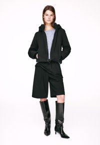 Čierna mikina na zips s kapucňou so s�ťahovacou šnúrkou, sivý top pod ňou, čierne široké krátke nohavice a lesklé čierne vysoké čižmy na jehlovom podpätku po kolená.