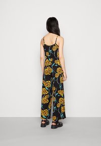 Černý overal s žlutým květinovým vzorem. Má tenké ramínka, přiléhavý pas a široké nohavice. Nosen s robustními černými botami.