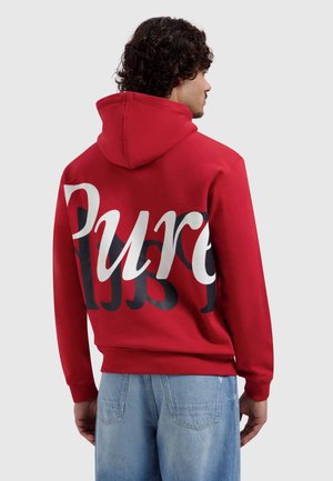 Man met een rode hoodie met de tekst "Pure" in grote witte en zwarte letters op de achterkant, gecombineerd met lichtblauwe spijkerbroek, met de rug naar de camera.