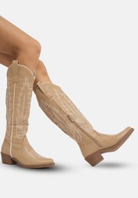 Botas de cuero marrón claro hasta la rodilla con patrones de relámpagos blancos cosidos, cierre con cremallera trasera y tacones cuadrados. Textura suave y diseño ajustado.