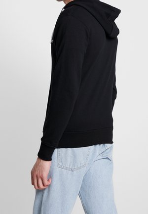 Mann trägt schwarzen Hoodie und hellblaue Jeans, seitlich und von hinten vor einfachem weißem Hintergrund gezeigt.