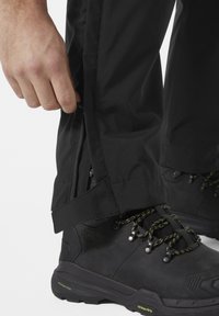 Zwarte waterdichte broek met ritssluitingen aan de onderkant, gedragen met zwarte wandelschoenen met groene accenten en een gestructureerde zool. Close-up van de details van de broek.