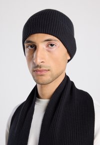 Musta ribattu neulottu pipo ja siihen sopiva huivi. Lähikuva, joka korostaa rakennetta ja muotoilua. Puettuna vaalean värisen paidan päälle.