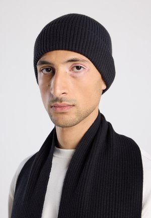 Zwarte geribbelde gebreide beanie en bijpassende sjaal. Close-up weergave die textuur en ontwerp benadrukt. Draagt boven een lichtgekleurde top.